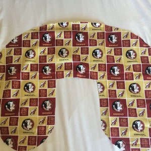 May include: A U-shaped nursing pillow with a red and yellow fabric featuring a repeated pattern of a Seminole mascot logo and the word "Seminoles".