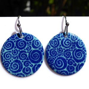 Op de afbeelding: Een paar ronde hangende oorbellen met een levendig patroon van donkerblauwe spiralen tegen een lichtblauwe achtergrond. Elke oorbel hangt aan een zilverkleurige metalen haak, wat zorgt voor een consistent en gebalanceerd ontwerp.