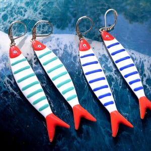 Può includere: Quattro paia di orecchini a forma di pesce. Gli orecchini hanno una base bianca con strisce orizzontali turchesi o blu. Ogni pesce ha una coda e una testa rosse. Sono esposti su una superficie blu a motivi di onde.