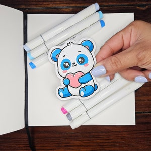 Puede incluir: Una pegatina de un panda azul y blanco con un corazón rosa. El panda está sosteniendo el corazón en sus patas. La pegatina está sobre una superficie blanca con marcadores azules y blancos.