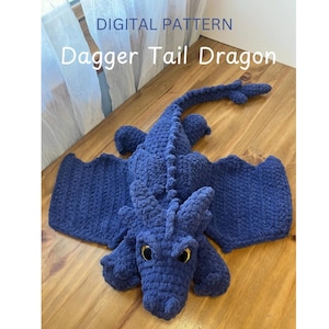 Könnte beinhalten: Ein gehäkelter blauer Drache mit einem Dolchschwanz, Flügeln und großen Augen. Der Drache liegt auf einer Holzoberfläche. Der Text "DIGITAL PATTERN Dagger Tail Dragon" befindet sich über dem Drachen.