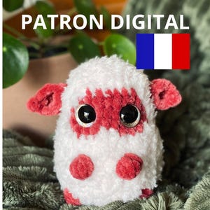 Può includere: Un giocattolo di peluche all'uncinetto a forma di mucca con pelliccia bianca e dettagli rossi. Il giocattolo ha grandi occhi neri e un naso rosso. Il giocattolo è seduto su una coperta verde. Il testo "PATRON DIGITAL" è visibile nell'immagine.