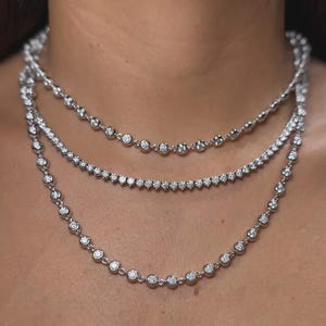 May include: A layered necklace set consisting of three distinct silver-toned chains. Two chains feature a series of evenly spaced, round, bezel-set clear stones, while the middle chain is a classic tennis-style necklace with closely set stones.