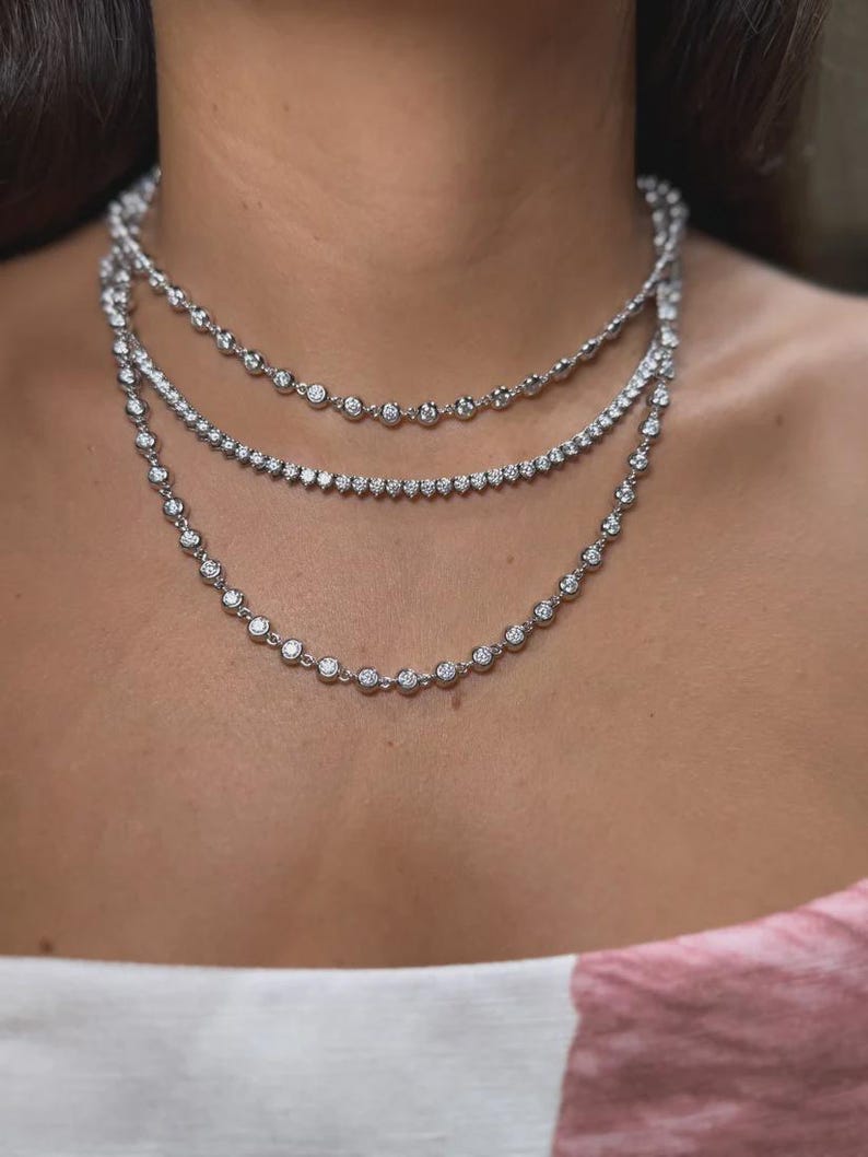 May include: A layered necklace set consisting of three distinct silver-toned chains. Two chains feature a series of evenly spaced, round, bezel-set clear stones, while the middle chain is a classic tennis-style necklace with closely set stones.