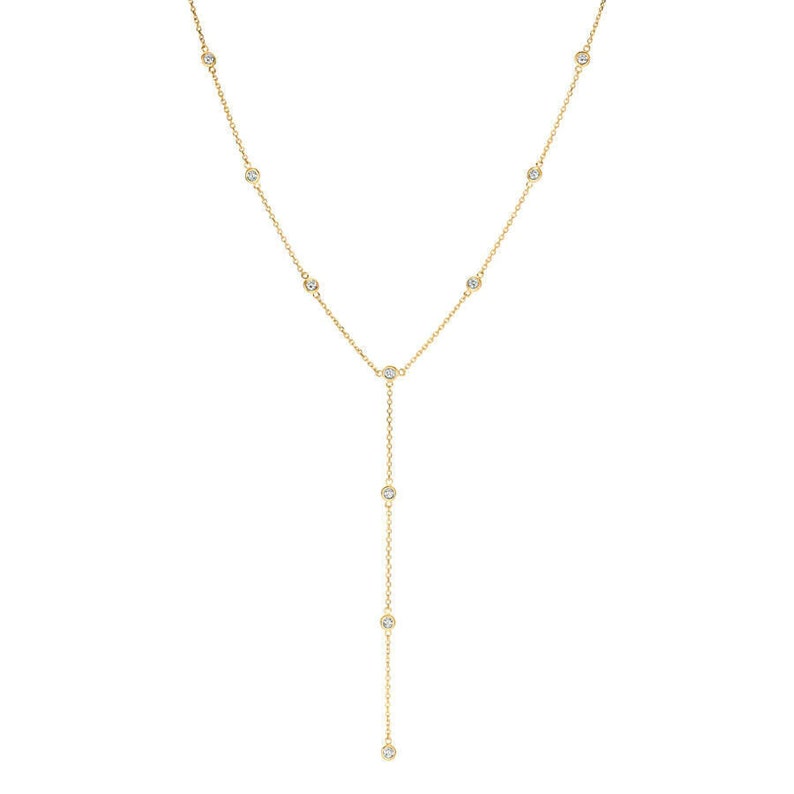 May include: A gold lariat necklace featuring a delicate chain with evenly spaced bezel-set round clear stones. The design includes a central junction point where the chain continues downward into a single vertical drop, accented with additional stones.
