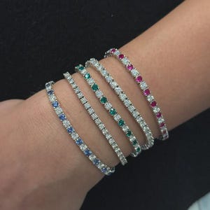 May include: A collection of six tennis bracelets, each featuring a row of gemstones. The bracelets display a variety of colors, including blue, green, and red, set against a silver or white metal. The bracelets are displayed on a person's wrist.
