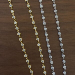 May include: Two delicate necklaces, one in a gold-tone metal and one in a silver-tone metal, each featuring a continuous chain of small, round, bezel-set clear stones that catch the light, arranged in a straight vertical line against a dark surface.