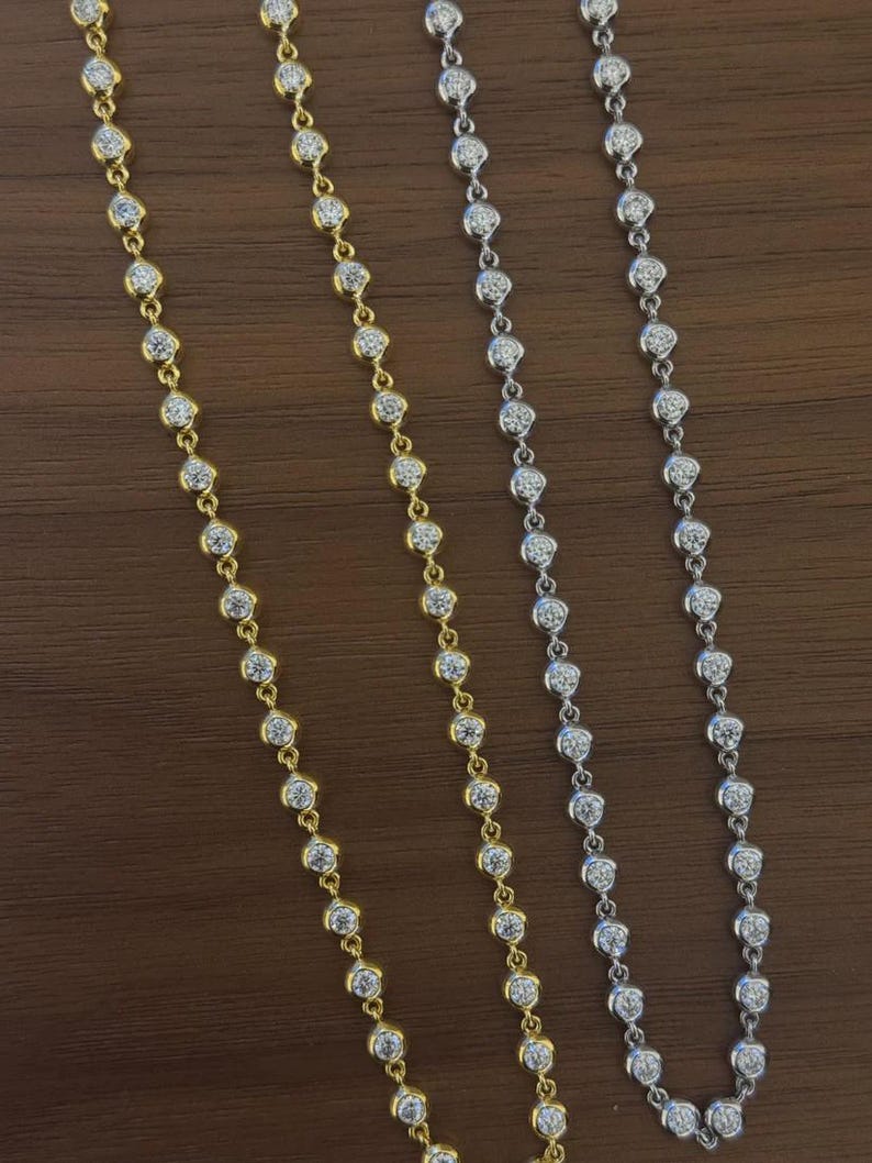 May include: Two delicate necklaces, one in a gold-tone metal and one in a silver-tone metal, each featuring a continuous chain of small, round, bezel-set clear stones that catch the light, arranged in a straight vertical line against a dark surface.