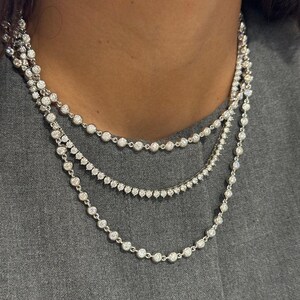 May include: A layered necklace set consisting of three separate silver-toned chains. Each chain features a series of small, round, clear stones set in individual metal bezels, creating a continuous sparkling effect around the neck.