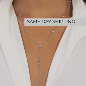 May include: A silver-toned Y-shaped lariat necklace featuring a delicate chain adorned with evenly spaced, round, clear bezel-set stones. The necklace design includes a vertical drop section that extends downward from the center point of the chain.