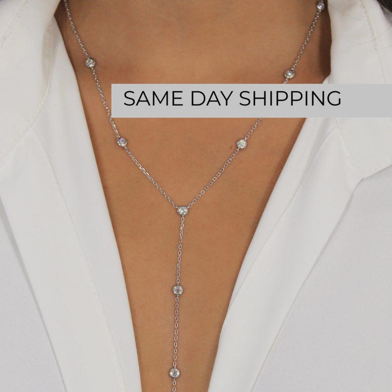 May include: A silver-toned Y-shaped lariat necklace featuring a delicate chain adorned with evenly spaced, round, clear bezel-set stones. The necklace design includes a vertical drop section that extends downward from the center point of the chain.