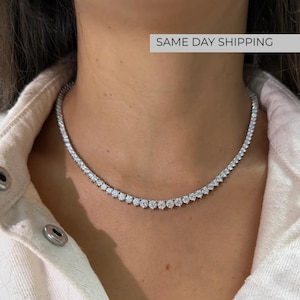 May include: A silver-toned tennis necklace featuring a continuous row of round, clear, faceted stones set in a metallic frame. The necklace rests against the neck, showcasing a uniform design of sparkling, circular gems linked together in a chain.