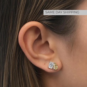 May include: A pair of stud earrings featuring two distinct gemstone shapes. One stone is a clear, rectangular emerald-cut gem, and the other is a yellow, pear-shaped teardrop gem. Both stones are set in a metallic frame and worn on an earlobe.