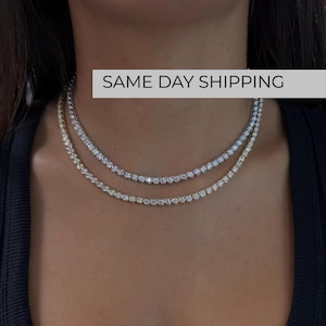 May include: Two layered tennis necklaces featuring round, clear, sparkling stones set in metal. One necklace is silver-toned, and the other is gold-toned. Both necklaces are worn around the neck, showcasing a classic, continuous link design.