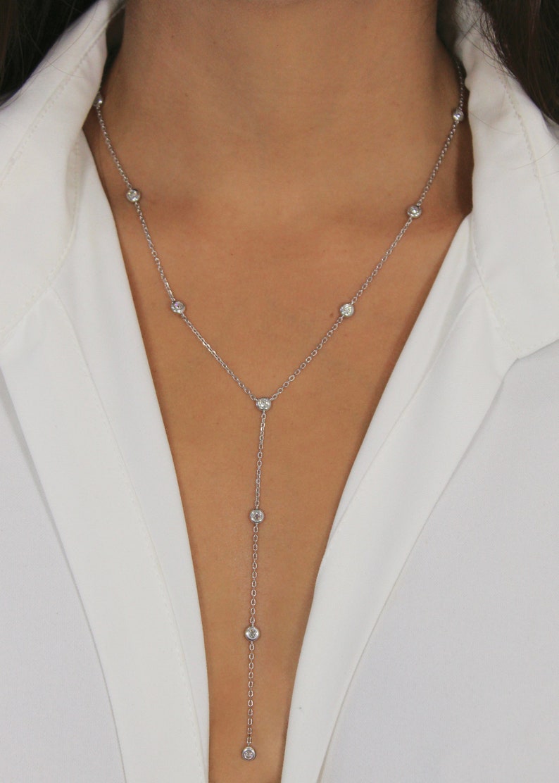May include: A silver-toned Y-shaped lariat necklace featuring a delicate chain with evenly spaced, round, clear bezel-set stones. The necklace drapes down the center of the chest, creating a vertical drop design with a single stone at the end.