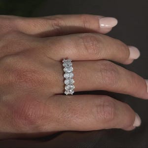 Pu&ograve; includere: Primo piano di una mano che indossa un anello d'argento. L'anello presenta una serie di pietre preziose chiare a taglio ovale, disposte in fila. Lo sfondo &egrave; sfocato e di tono neutro.
