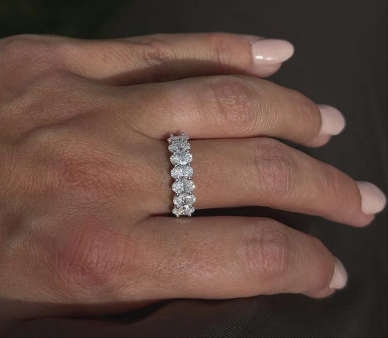 Pu&ograve; includere: Primo piano di una mano che indossa un anello d'argento. L'anello presenta una serie di pietre preziose chiare a taglio ovale, disposte in fila. Lo sfondo &egrave; sfocato e di tono neutro.