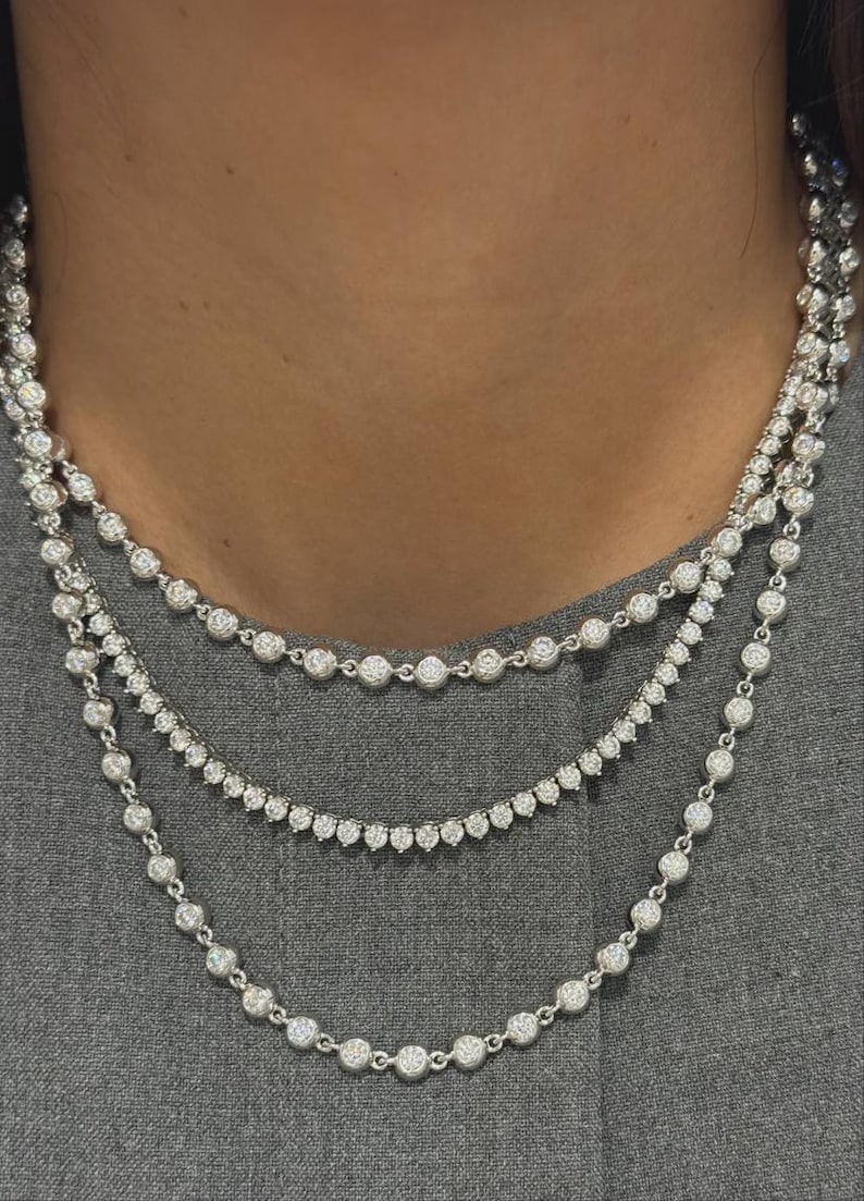 May include: A layered necklace set consisting of three separate silver-toned chains. Each chain features a series of round, clear, sparkling stones set in individual metal bezels, creating a continuous, uniform, and reflective design around the neck.