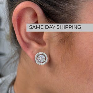 May include: A circular stud earring featuring a large, round, clear faceted centre stone surrounded by a halo of smaller, sparkling clear stones set in a polished silver-toned metal frame, securely fastened to the earlobe.