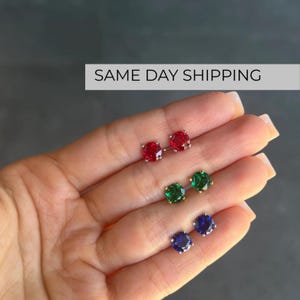 Può includere: Tre paia di orecchini a lobo rotondi con pietre preziose sfaccettate in rosso, verde e blu. Ogni orecchino è montato su una struttura metallica a griffe, con un design a taglio brillante che riflette la luce sulla superficie delle pietre.