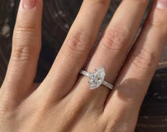 Pode incluir: Um anel de noivado com diamante de corte marquesa e uma banda prateada. O anel apresenta um grande diamante transparente em forma de marquesa como peça central, com diamantes menores incrustados ao longo da banda. O anel é mostrado numa mão com unhas cuidadas.