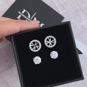 May include: A black box with the "Diamonds" logo displays two pairs of diamond earrings. One pair has a halo design, the other is a classic round cut. The earrings are set in silver or white gold.