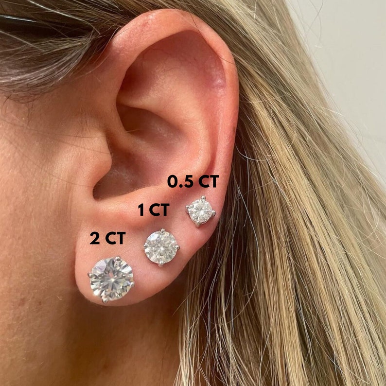 May include: Close-up of a person's ear with three different sizes of round, clear, cubic zirconia earrings. The earrings are labeled 0.5 CT, 1 CT, and 2 CT.