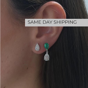 May include: A pair of silver-toned earrings consisting of one pear-shaped clear stone stud and one drop earring featuring a pear-shaped green stone at the top and a pear-shaped clear stone dangling below, set in a metallic frame.