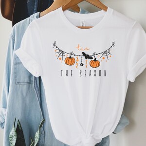 May include: White t-shirt with a black and orange graphic of a Halloween garland with pumpkins, bats, and stars. The text "Tis the Season" is printed below the garland.