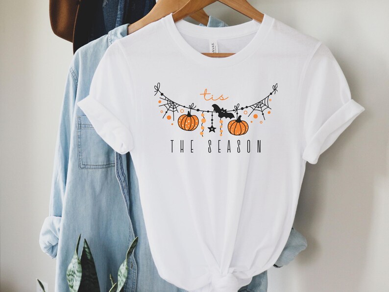 May include: White t-shirt with a black and orange graphic of a Halloween garland with pumpkins, bats, and stars. The text "Tis the Season" is printed below the garland.