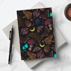 May include: A black notebook with a repeating pattern of mushrooms, butterflies, frogs, pine cones, and crescent moons. The pattern is in shades of red, purple, blue, green, and yellow.