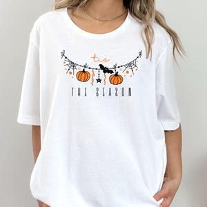May include: White t-shirt with a graphic of a Halloween garland with pumpkins, bats, and stars. The text "tis the season" is printed below the garland.