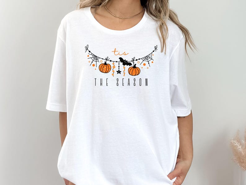 May include: White t-shirt with a graphic of a Halloween garland with pumpkins, bats, and stars. The text "tis the season" is printed below the garland.