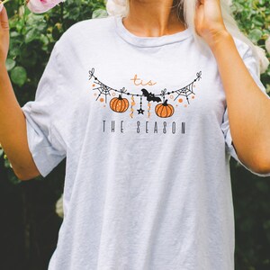 May include: A white t-shirt with a graphic of a string of pumpkins, bats, and spiderwebs. The text "Tis the Season" is printed below the graphic.