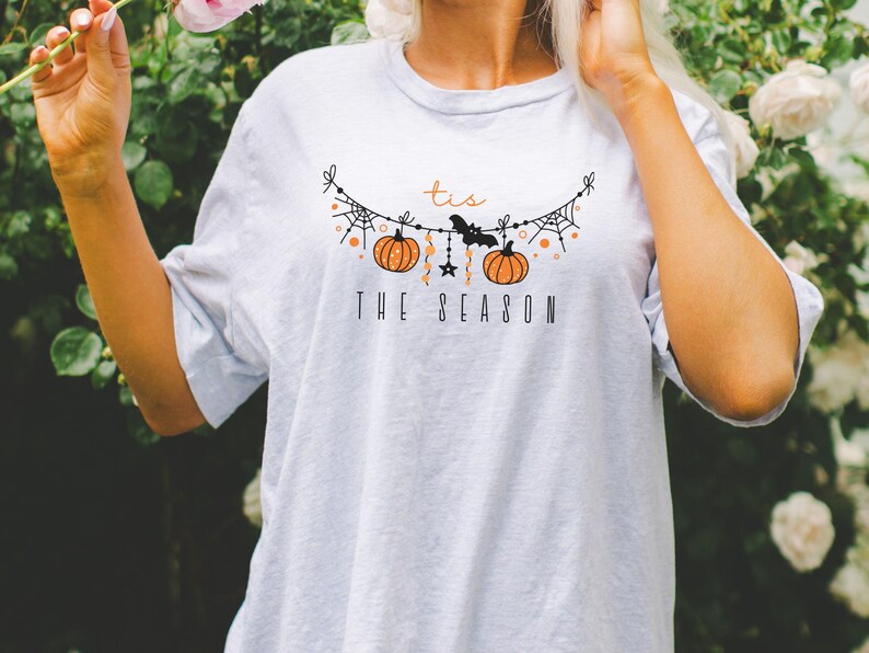 May include: A white t-shirt with a graphic of a string of pumpkins, bats, and spiderwebs. The text "Tis the Season" is printed below the graphic.