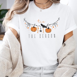 May include: White t-shirt with a black and orange graphic of a Halloween garland with pumpkins, bats, and spiderwebs. The text "'tis the season" is printed below the garland.