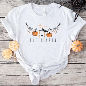 May include: A white t-shirt with a black and orange graphic design. The graphic features a string of pumpkins, bats, and spiderwebs with the text "'tis the season".