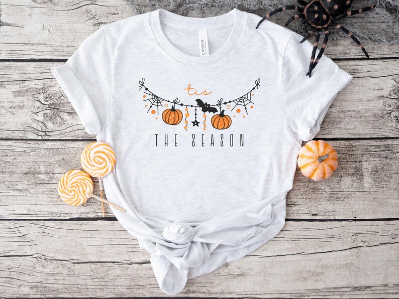 May include: A white t-shirt with a black and orange graphic design. The graphic features a string of pumpkins, bats, and spiderwebs with the text "'tis the season".