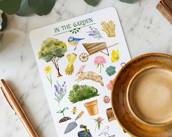 May include: A sheet of garden-themed stickers with watercolor illustrations of a tree, bunny, wheelbarrow, flowers, and gardening tools. The text "IN THE GARDEN" is at the top. A cup of coffee and a pen are in the foreground.