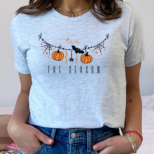 May include: A gray t-shirt with a graphic of a Halloween garland with pumpkins, bats, and stars. The text "Tis the Season" is printed below the garland.