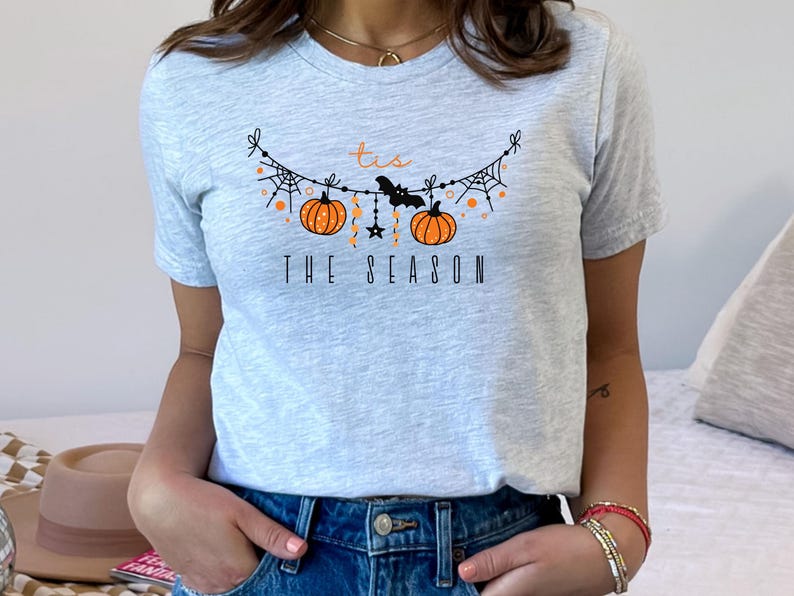 May include: A gray t-shirt with a graphic of a Halloween garland with pumpkins, bats, and stars. The text "Tis the Season" is printed below the garland.