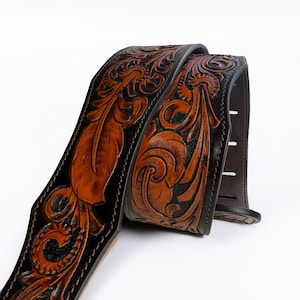 May include: A brown and black leather guitar strap with intricate floral and leaf carvings. The strap features a detailed, embossed design with a dark brown border and stitching. The guitar strap is a musical instrument accessory.