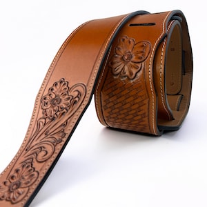 Pode incluir: Uma correia de guitarra de couro castanho enrolada com detalhes florais e entrançados. A correia apresenta desenhos de flores esculpidas e costuras brancas. A correia tem aproximadamente 7.6 cm de largura.