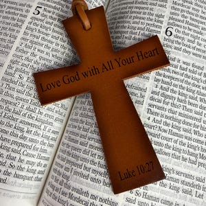 Könnte beinhalten: Ein braunes ledernes kreuzförmiges Lesezeichen mit dem Text "Love God with All Your Heart" und "Luke 10:27" aufgedruckt. Das Lesezeichen liegt auf einem offenen Buch.
