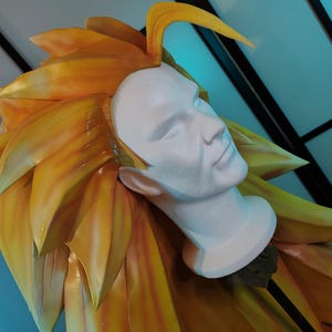 Puede incluir: Una cabeza de maniquí blanca con un rostro y cuello detallados está rodeada de cabello grande, puntiagudo, amarillo y naranja. El cabello está peinado para que se levante y sobresalga, creando un efecto dramático. El fondo es azul claro.