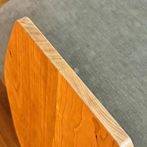 May include: Close-up of a wooden object with a light brown, polished surface. The object has a rounded edge and a natural wood grain pattern. The edge is a lighter color, and the background is a gray fabric.