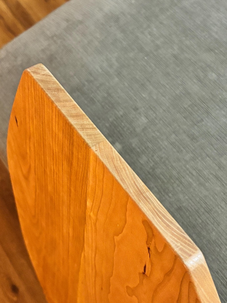 May include: Close-up of a wooden object with a light brown, polished surface. The object has a rounded edge and a natural wood grain pattern. The edge is a lighter color, and the background is a gray fabric.