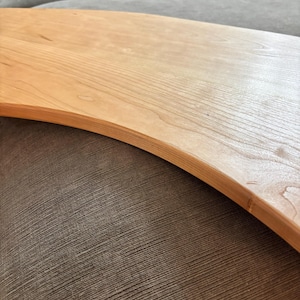 May include: A light-colored wooden serving board with a curved edge. The wood grain is visible, and the board appears to be smooth. The board is resting on a textured, brown surface.