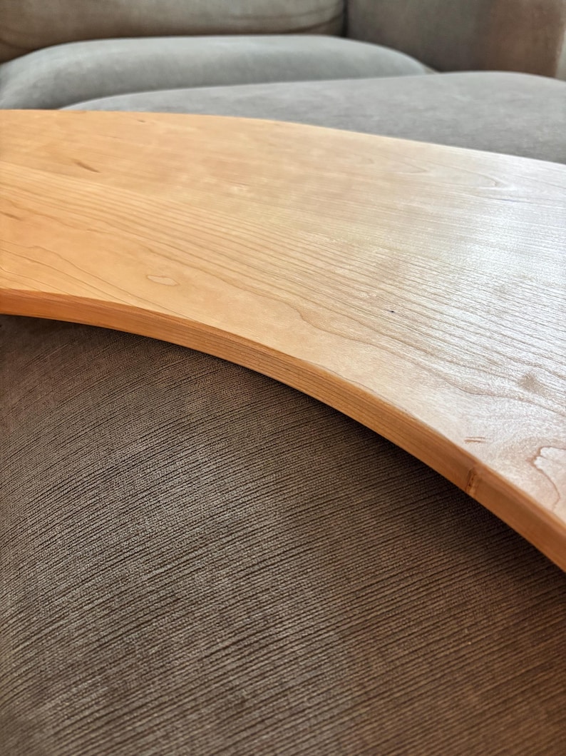 May include: A light-colored wooden serving board with a curved edge. The wood grain is visible, and the board appears to be smooth. The board is resting on a textured, brown surface.