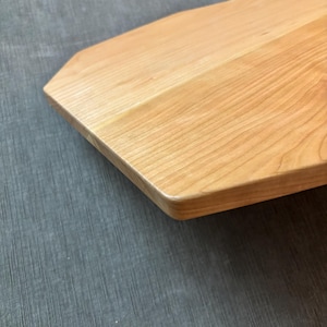May include: Close-up of a light-colored wooden cutting board with a unique, angled shape. The wood grain is visible, and the edges are rounded. The board is likely used for food preparation and serving.